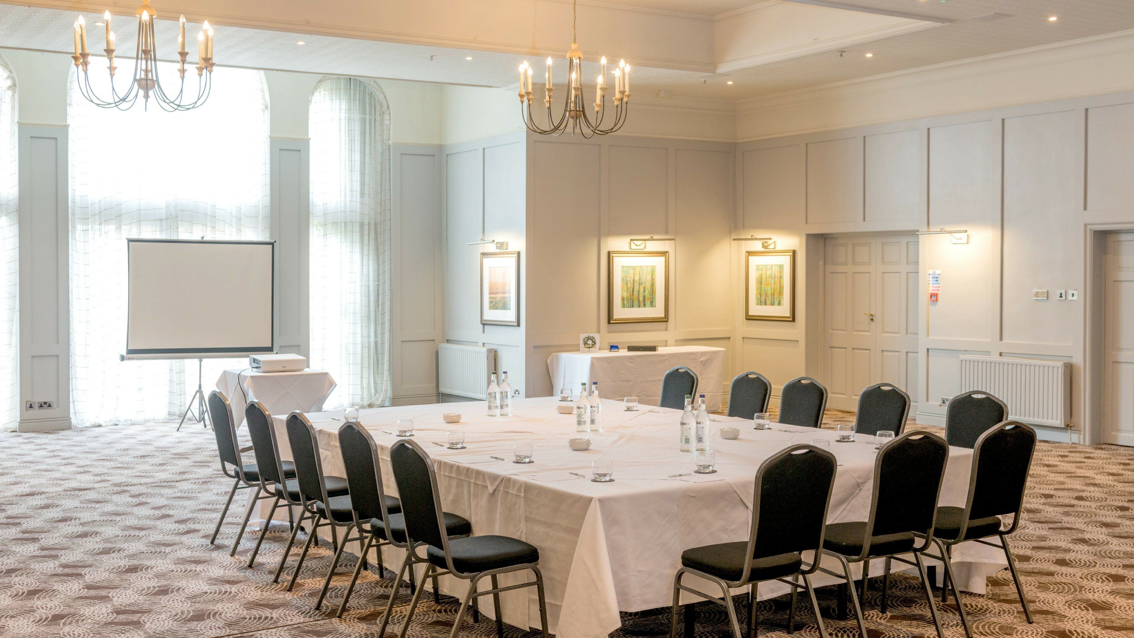 A conference room with a long table, chairs, a projector screen, and framed artwork on the walls.