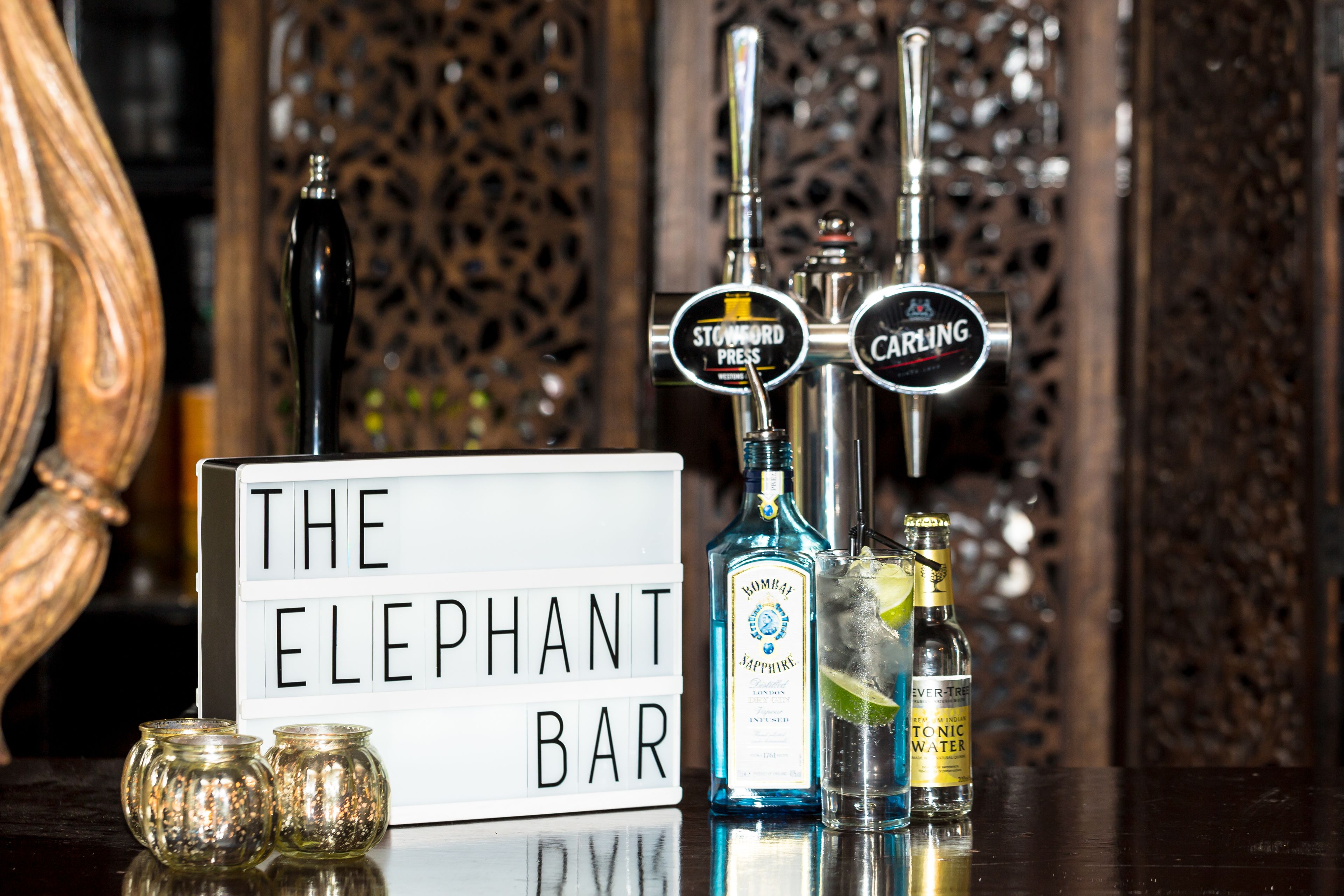 A bar setup with a sign reading "The Elephant Bar," bottles of Bombay Sapphire gin, and a bottle of tonic water.