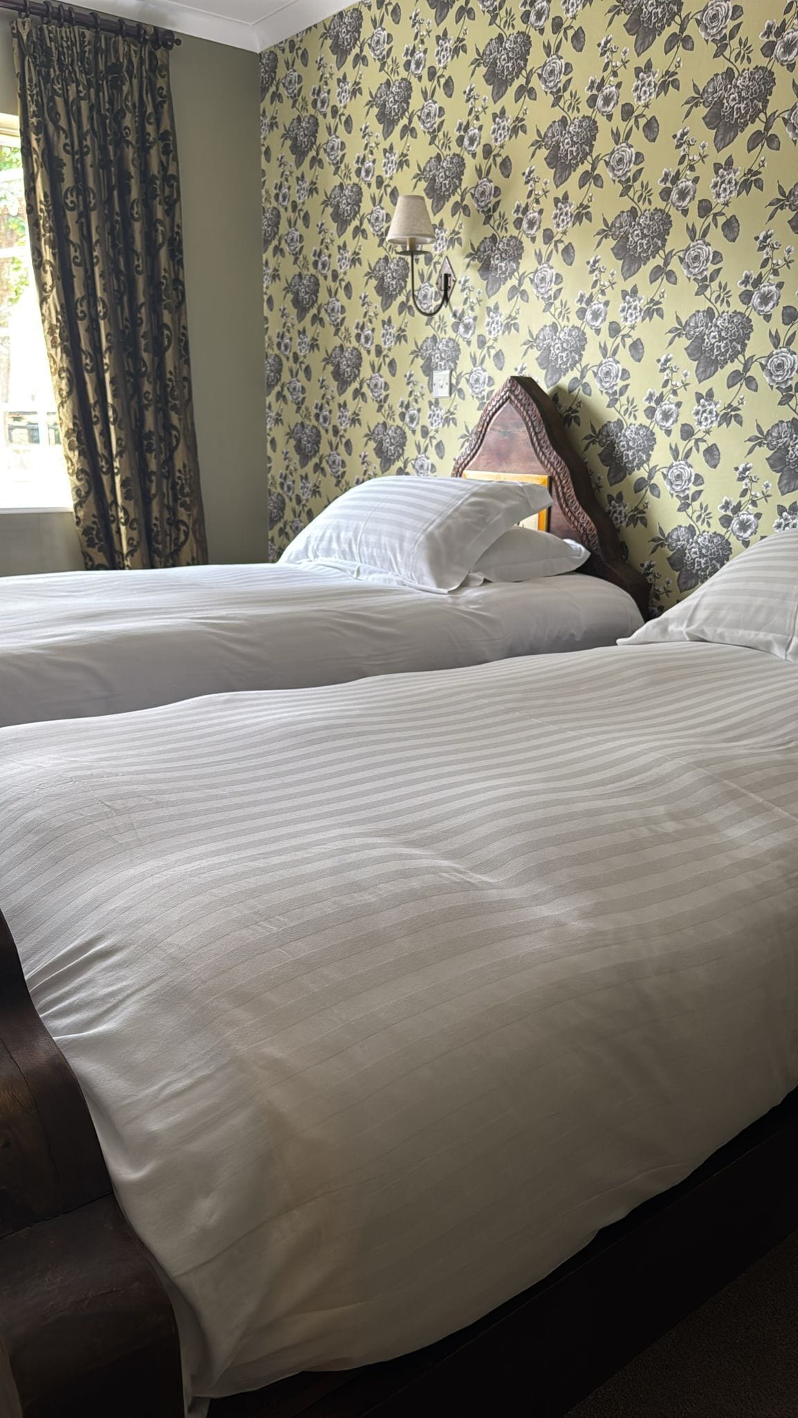Et hotelværelse med to senge, blomstret tapet, en lampe og et vindue med gardiner.