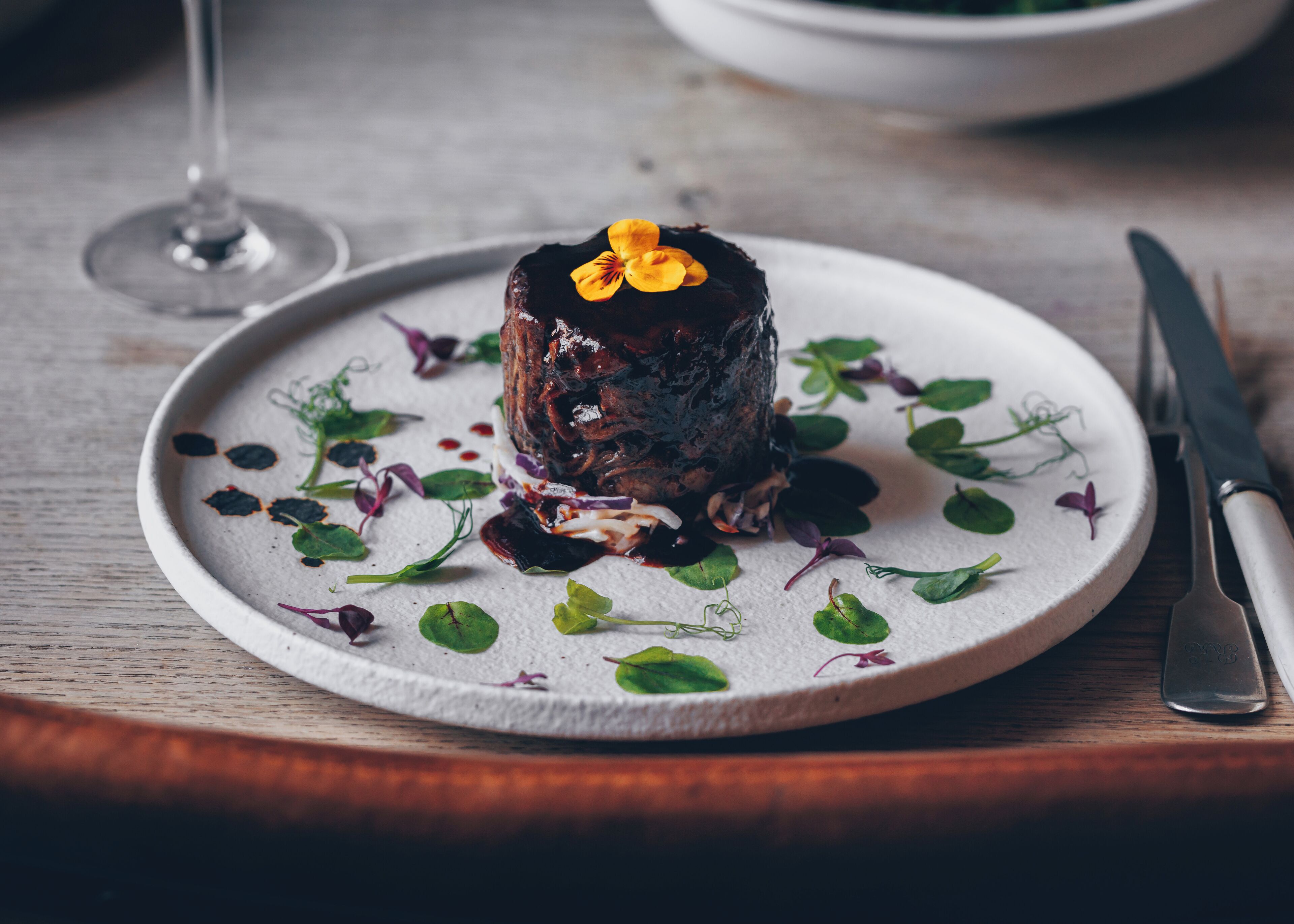A plate with a meat dish, garnished with greens and a flower, on a wooden table.