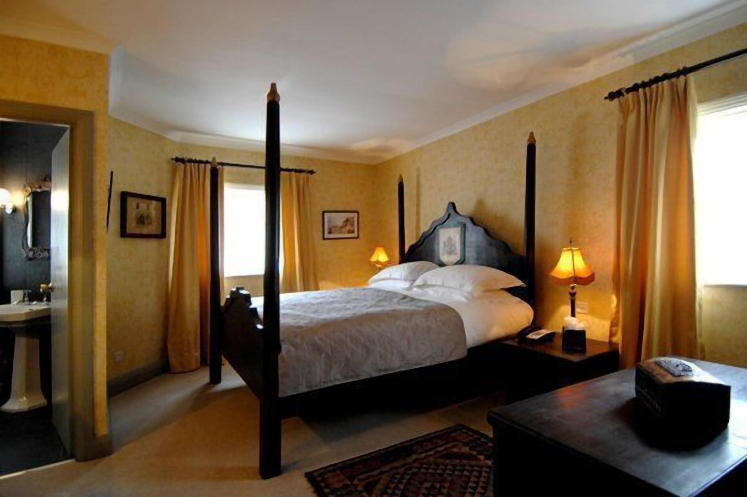 A four-poster bed with a headboard, two bedside lamps, and a nightstand with a lamp. A small table is beside the bed, and a mirror is on the wall.