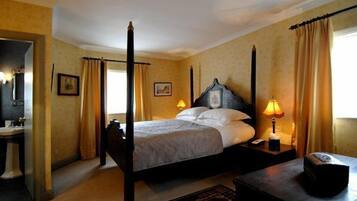 A four-poster bed with a headboard, two bedside lamps, and a nightstand with a lamp. A small table is beside the bed, and a mirror is on the wall.