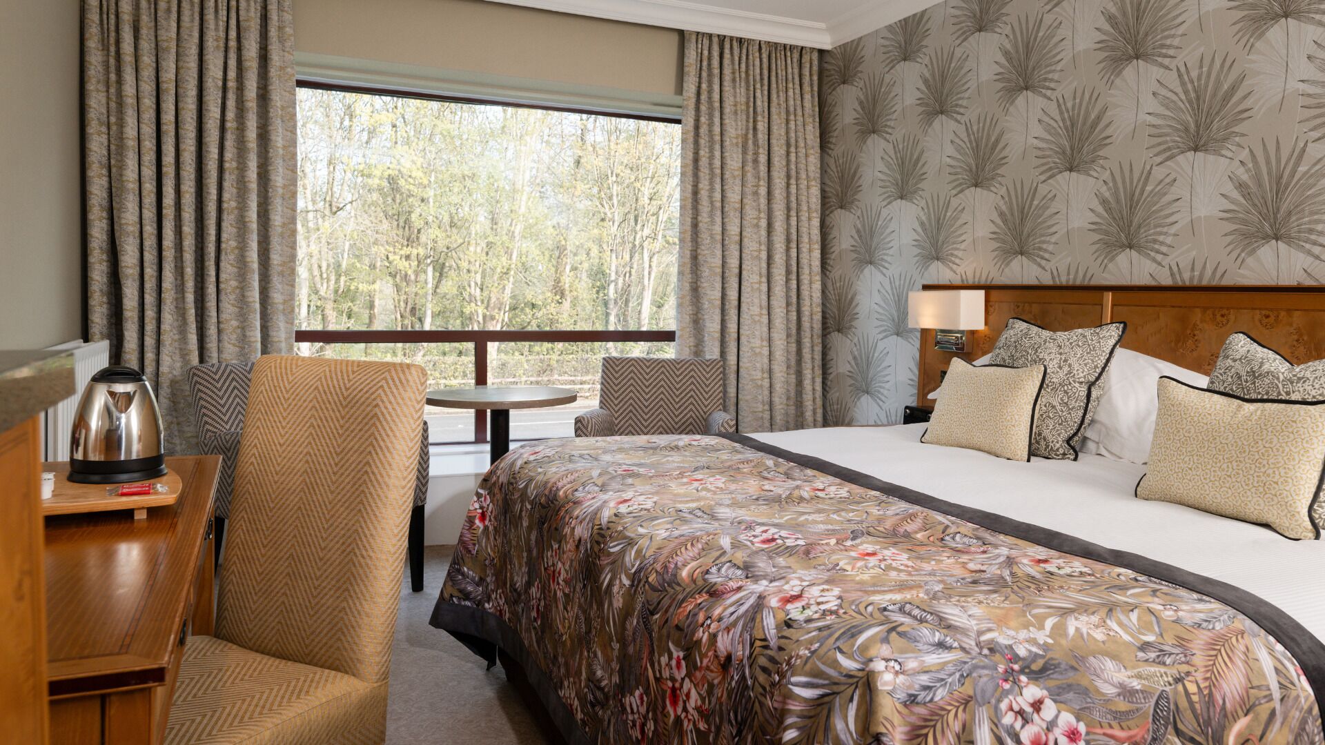 A hotel room with a large bed, a wooden desk, a chair, and a view of trees through the window.