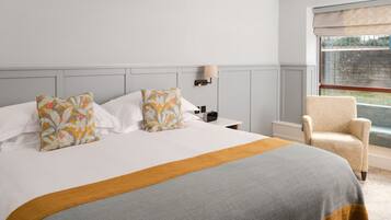 A neatly made bed with floral pillows, a bedside table with a lamp, and a chair by a window.