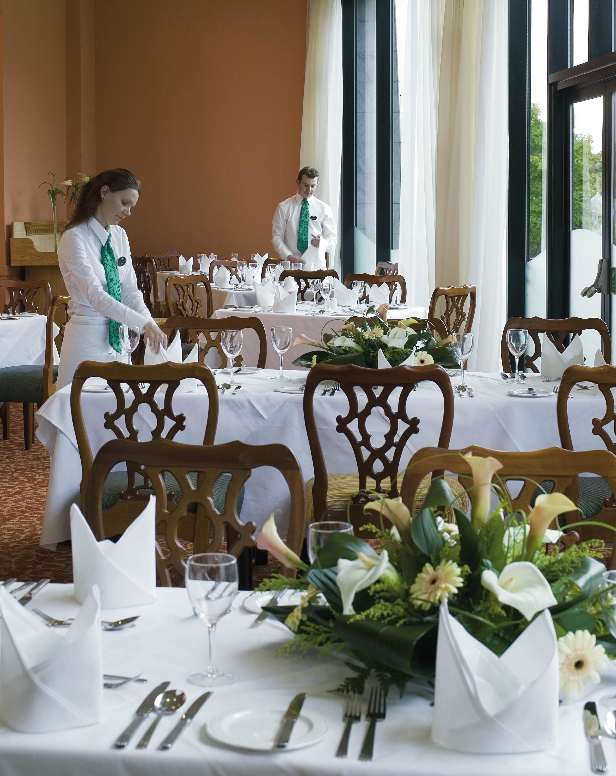 Un comedor con mesas dispuestas para una comida, con manteles blancos, sillas de madera y centros de mesa con flores.