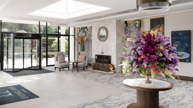 A modern hotel lobby with a glass entrance, seating area, fireplace, and a large floral centerpiece. - Everglades Hotel (Londonderry)