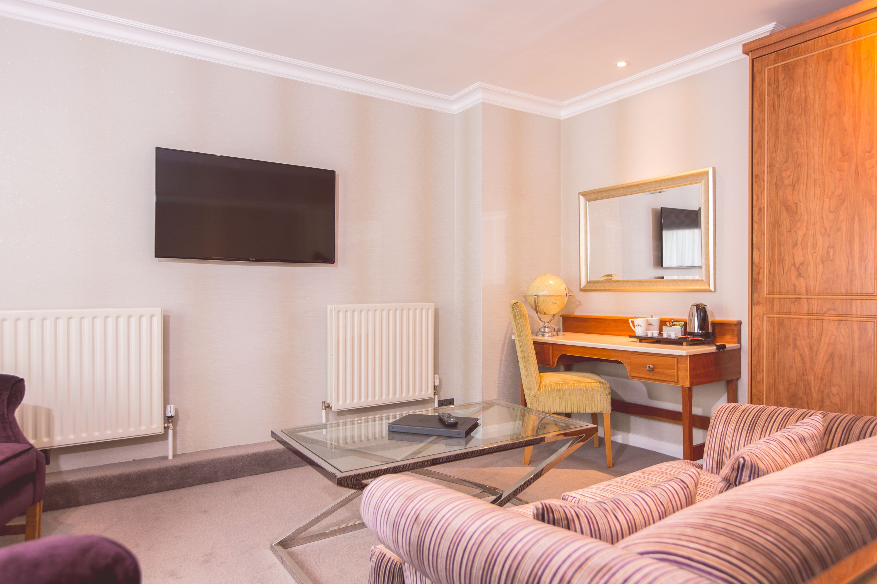 Habitación de hotel con sofá, una mesita de centro de vidrio, escritorio con silla, televisor de pantalla plana, radiador y armario de madera.