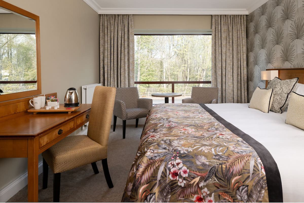A hotel room with a bed, a desk with a kettle, a chair, a table, and a view of trees through the window.
