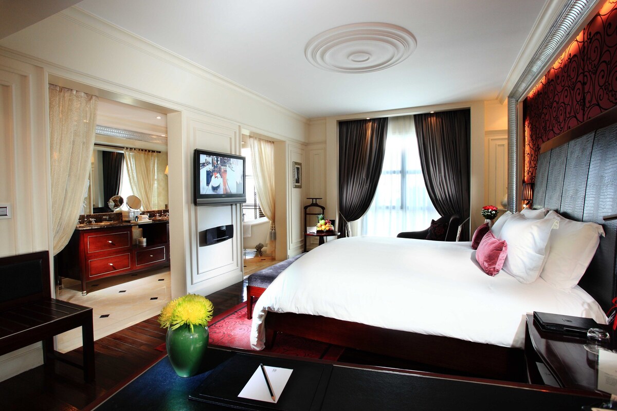 Ein Hotelzimmer mit einem großen Bett, einem Fernseher, einem Schreibtisch mit Stuhl, einem Schminktisch und einer Vase mit Blumen.