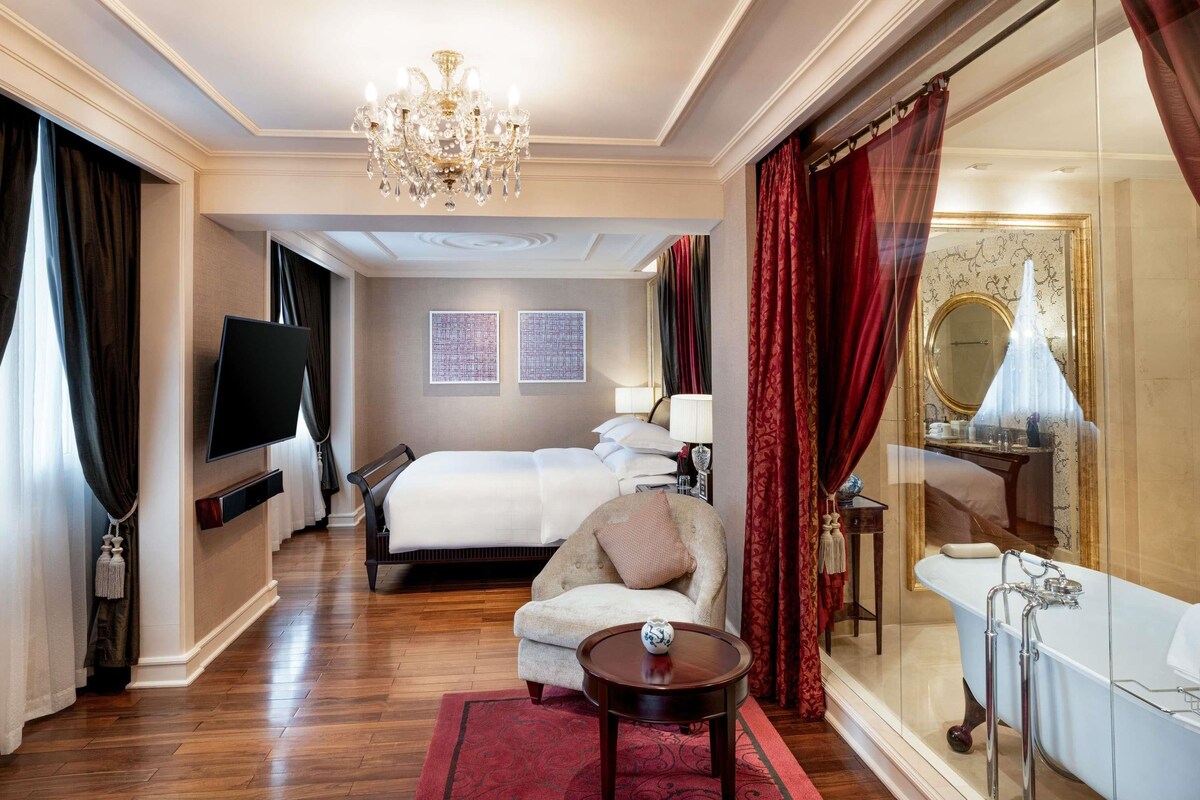 A hotel room with a bed, a sofa, a TV, a chandelier, a bathroom with a bathtub, and a large mirror.