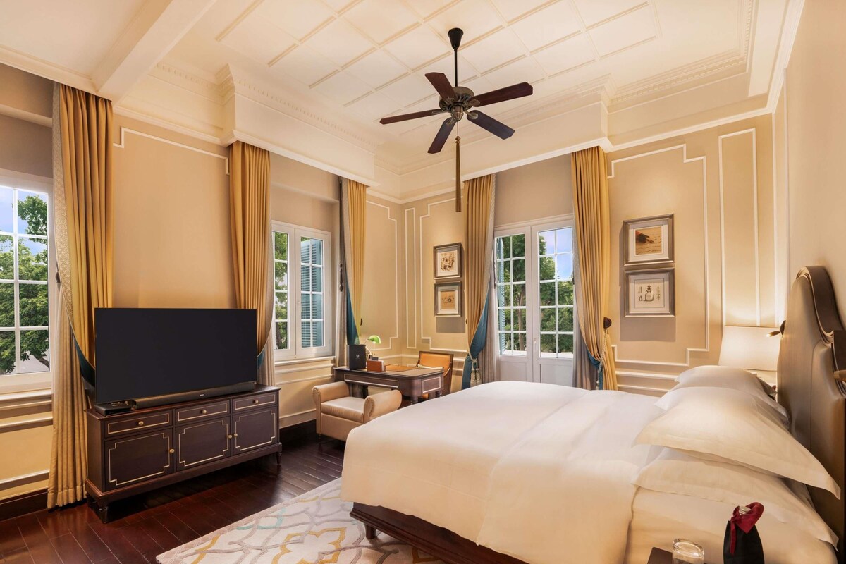 A hotel room with a large bed, a television, a desk with a chair, and a ceiling fan.