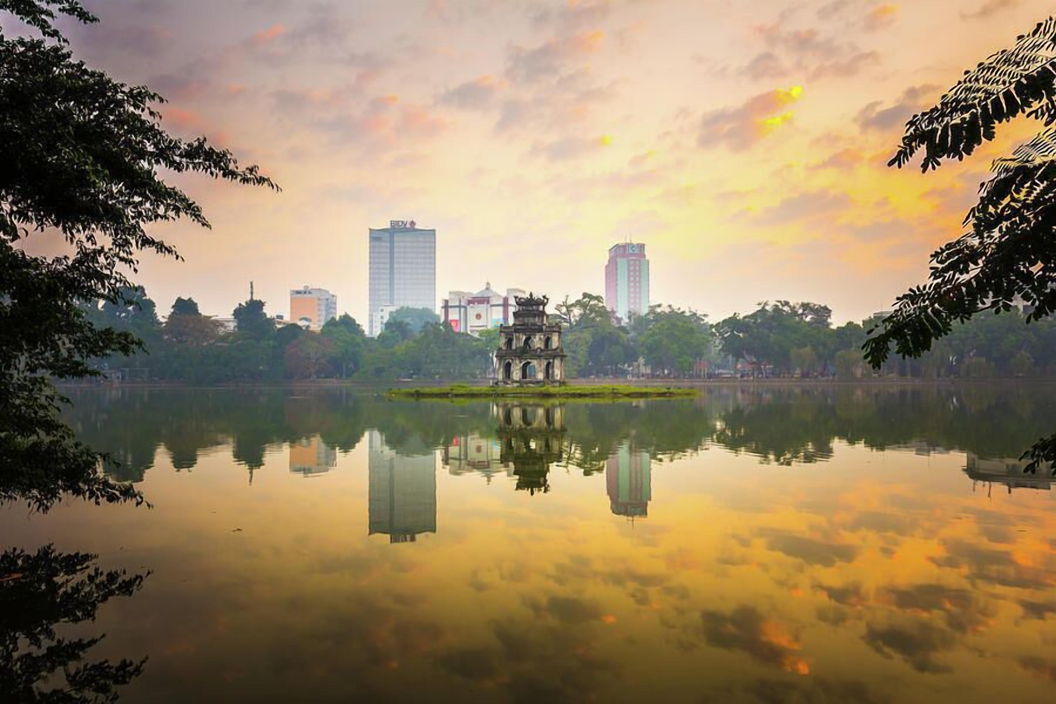 A serene lake with a traditional pavilion in the center, surrounded by modern buildings and lush greenery.