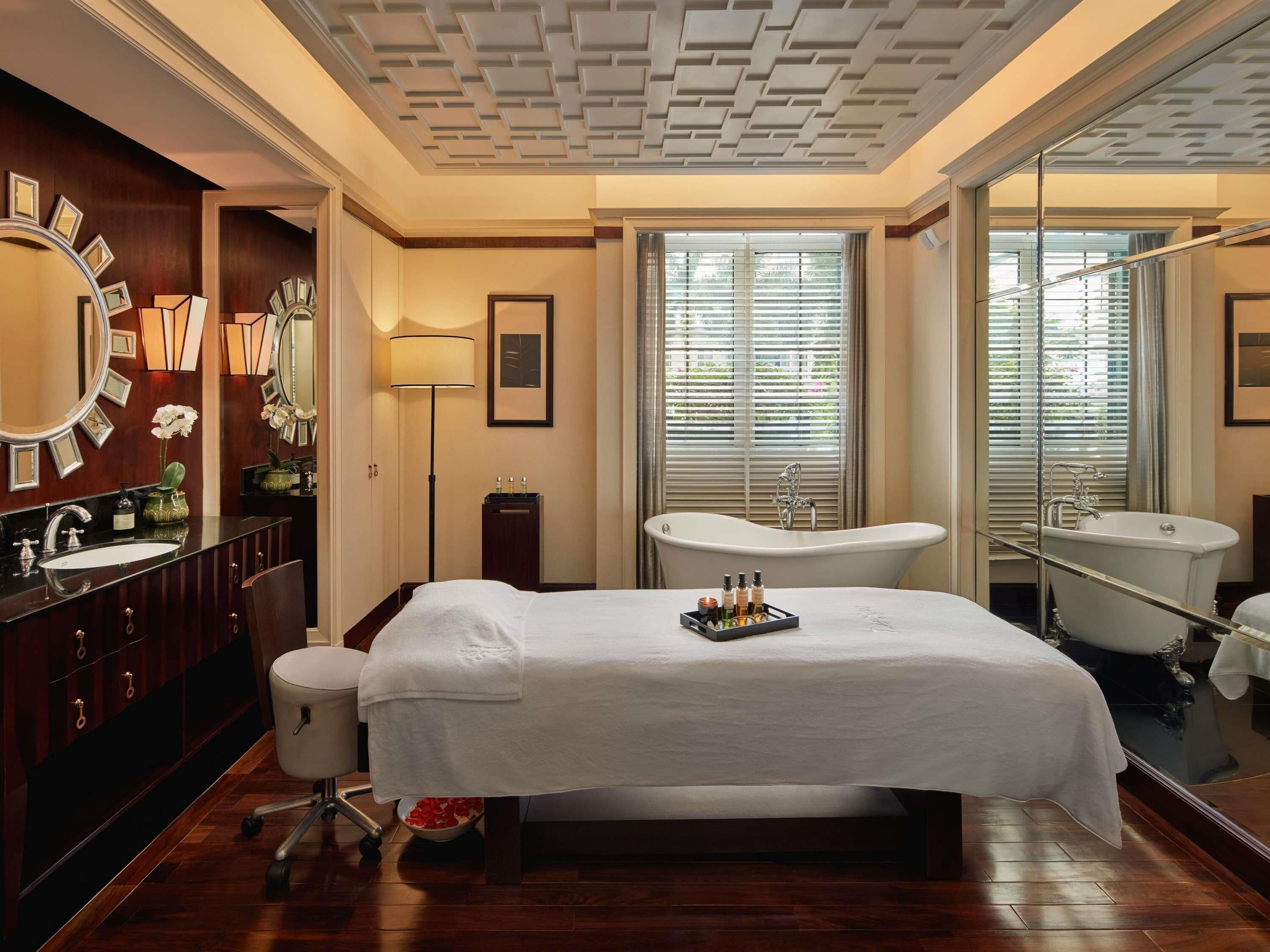 A hotel room with a large bed, a vanity with a mirror, a bathtub, and a large window with blinds.