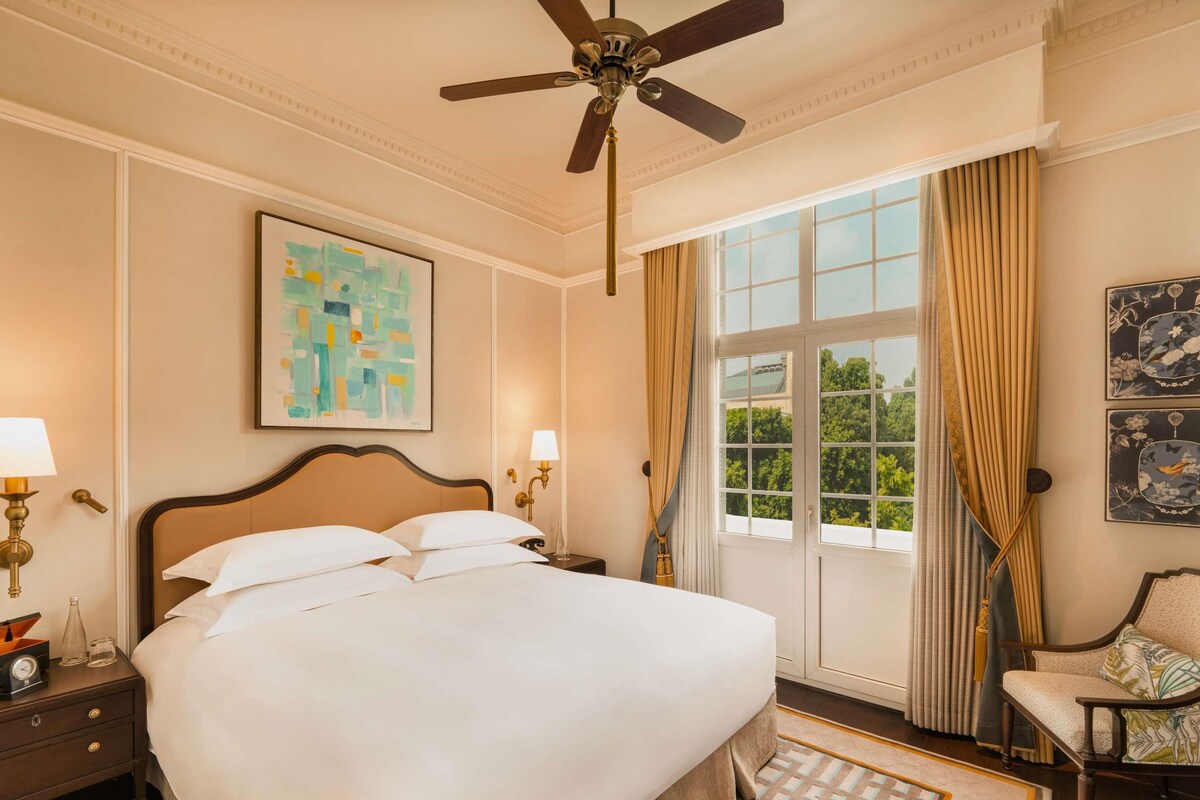 A hotel room with a large bed, a ceiling fan, a window with curtains, and artwork on the walls.