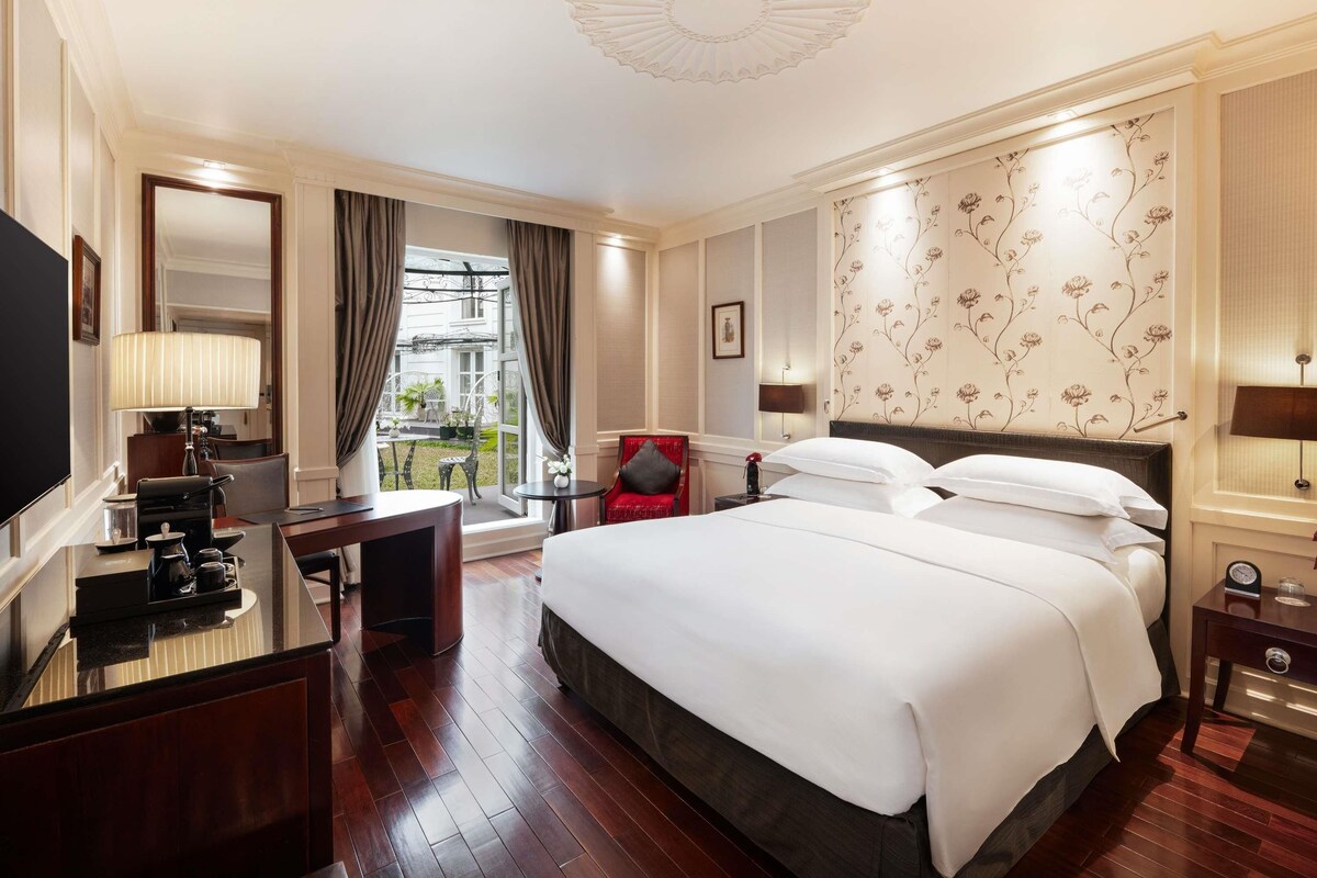 A hotel room with a large bed, a desk with a lamp, a TV, and a view of the outside.