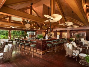 A spacious bar area with high ceilings and wooden beams, featuring a bar counter, multiple seating options including bar stools and wicker chairs, and ceiling fans. - Sofitel Legend Metropole Hanoi (Hanoi)
