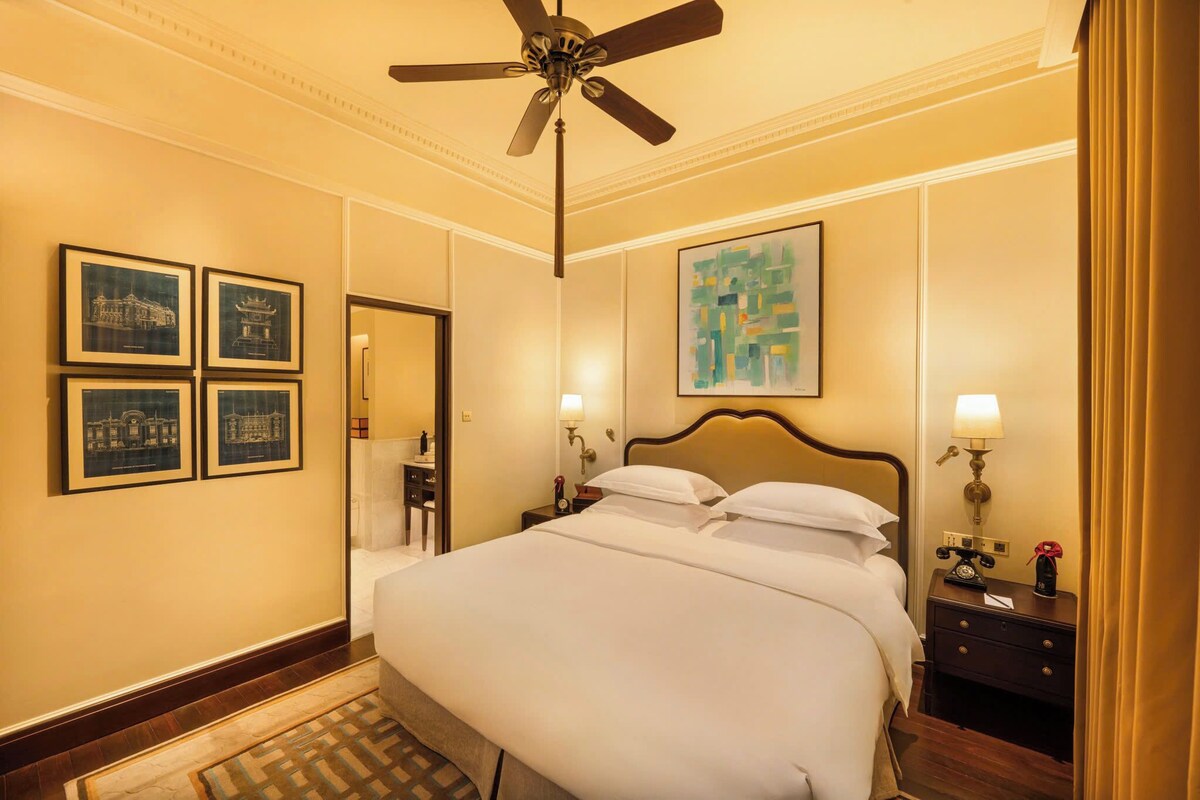 Ein Hotelzimmer mit einem großen Bett, einem Deckenventilator, gerahmten Kunstwerken an den Wänden, einem Nachttisch mit Lampe und einem Teppich auf dem Boden.