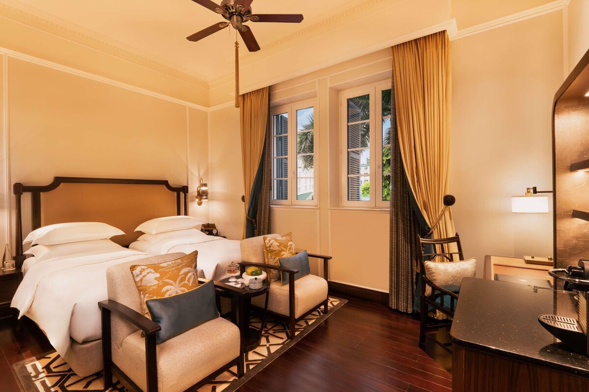 A hotel room with a bed, two armchairs, a small table, a desk, and a ceiling fan.