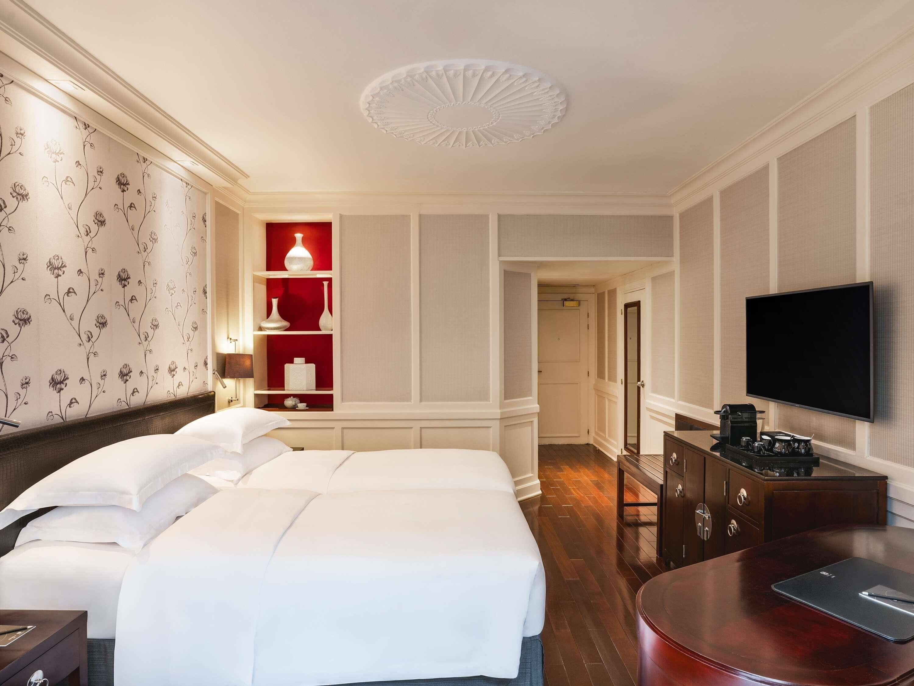 Quarto de hotel com uma cama grande, uma televisão, uma escrivaninha com um laptop, uma parede de destaque vermelha e um corredor que leva a outro quarto.