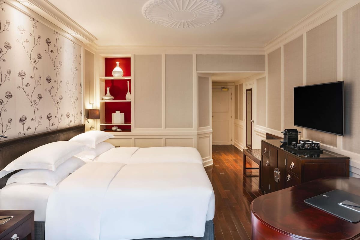 A hotel room with a large bed, a TV, a desk with a laptop, a red accent wall, and a hallway leading to another room.