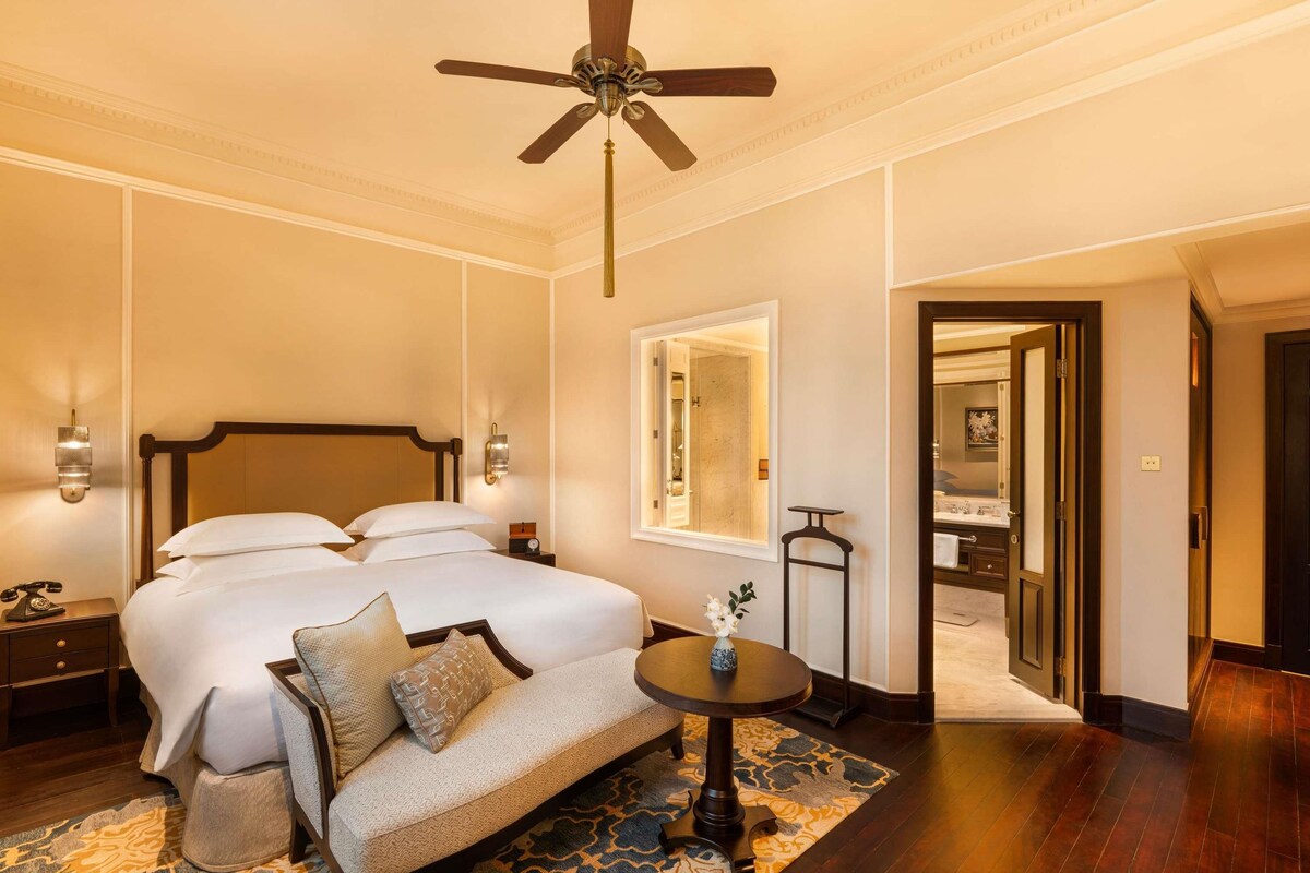 A hotel room with a large bed, bedside tables, a sofa, a small table with a vase, a ceiling fan, and a mirror.
