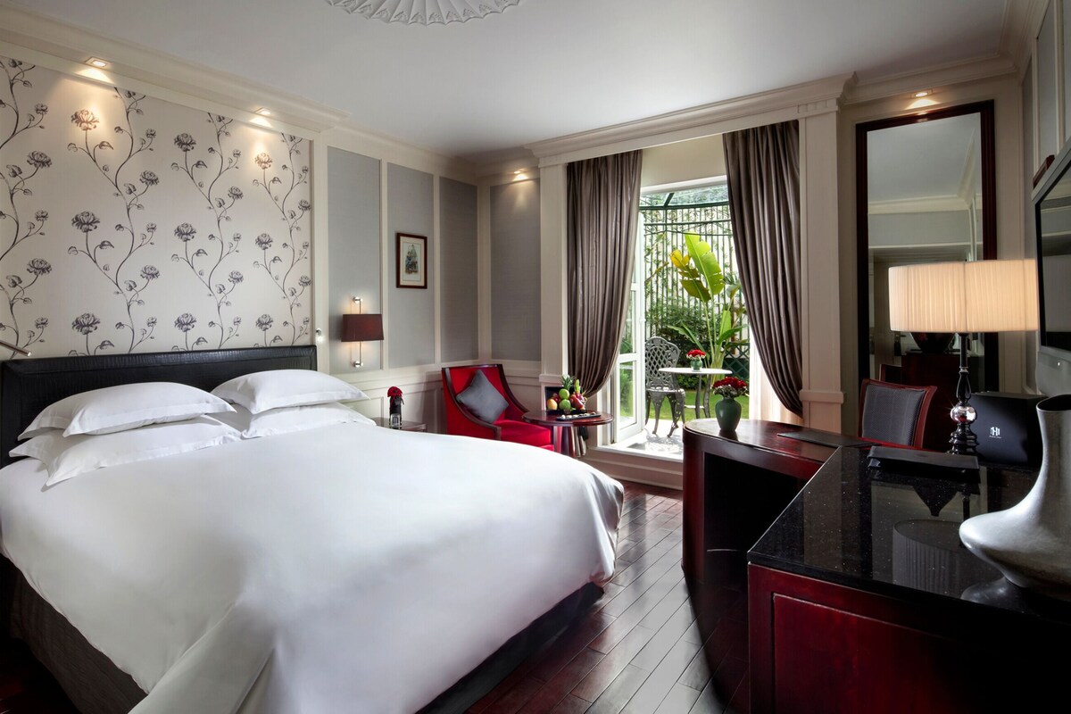 Ein Hotelzimmer mit einem großen Bett, einem Schreibtisch mit Lampe, einem Sessel und Blick auf Grün durch das Fenster.
