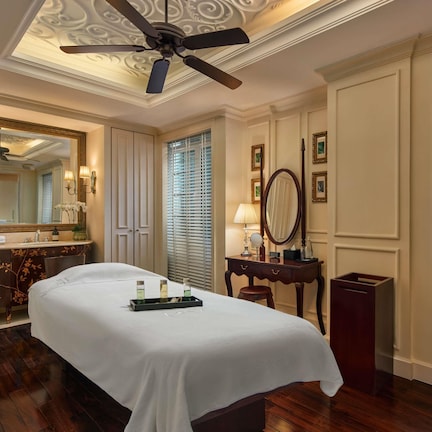 A hotel room with a bed, a vanity with a mirror, a ceiling fan, and a window with blinds.