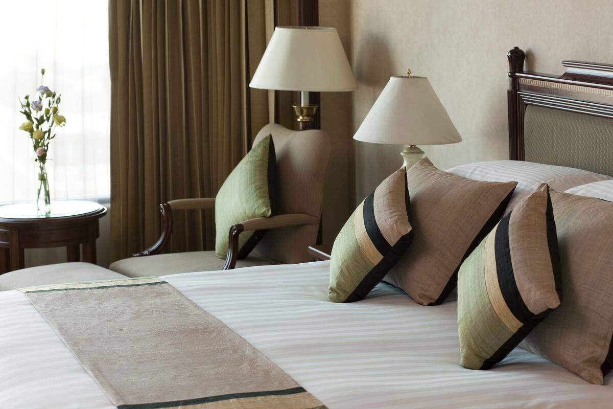 A hotel room with a bed, two lamps, a chair, a table with a vase of flowers, and a window with curtains.