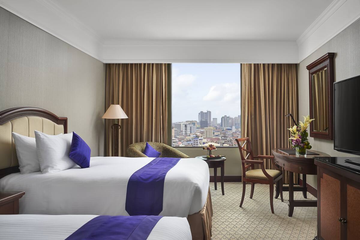 A hotel room with two beds, a desk, a chair, a TV, and a view of the cityscape.