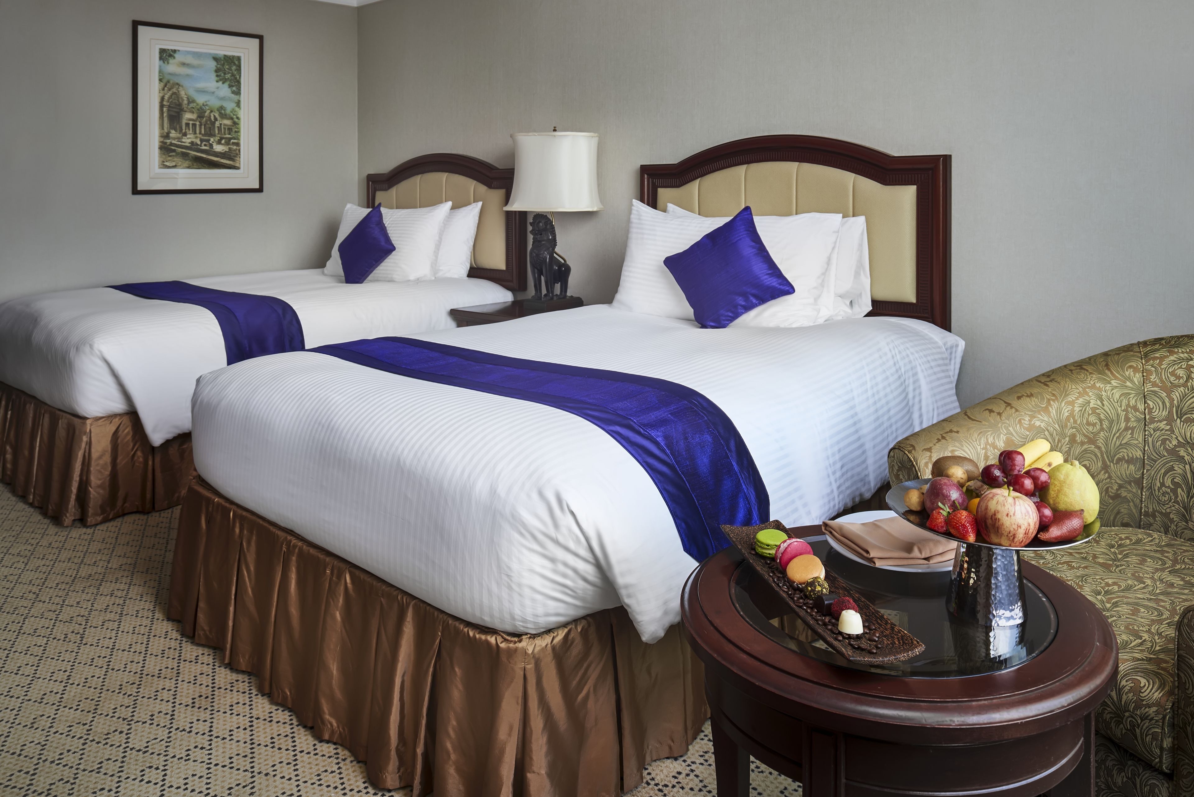 Une chambre d’hôtel avec deux lits, une table de chevet avec une lampe, un panier de fruits et un tableau encadré au mur.