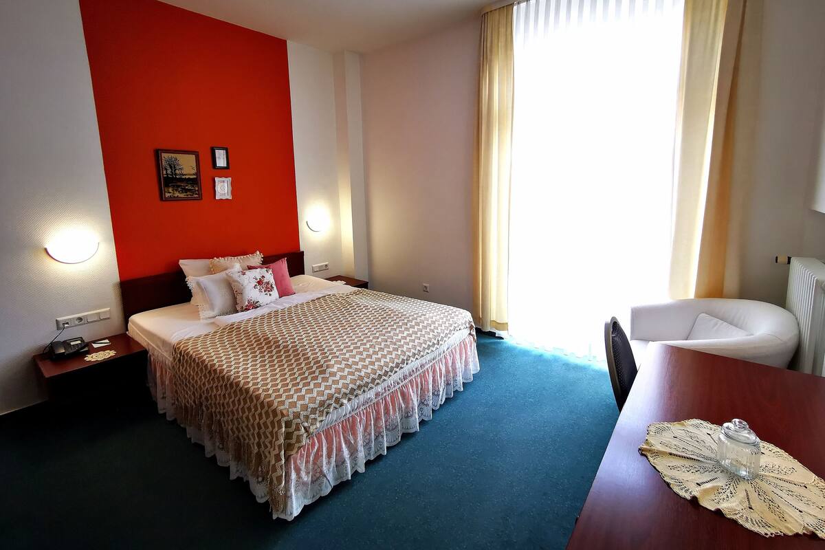 A hotel room with a bed, bedside table, chair, and a view of the outdoors.