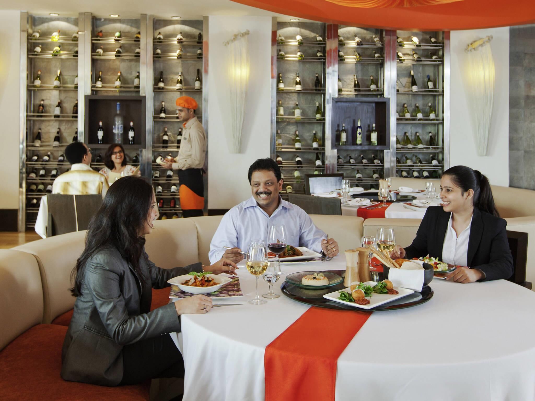 A restaurant with round tables, wine bottles on shelves, and a waiter in the background.