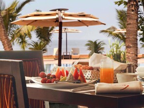 Une table dressée pour le petit-déjeuner, avec des assiettes de fruits, des croissants et un verre de jus d’orange, donnant sur une piscine et des palmiers.