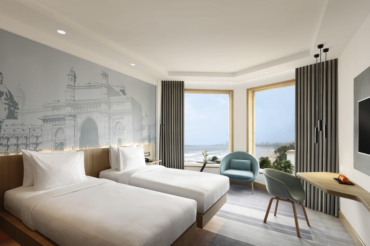 Ein Hotelzimmer mit zwei Betten, einem Schreibtisch, einem Stuhl, einem Fernseher und einem großen Fenster mit Meerblick.