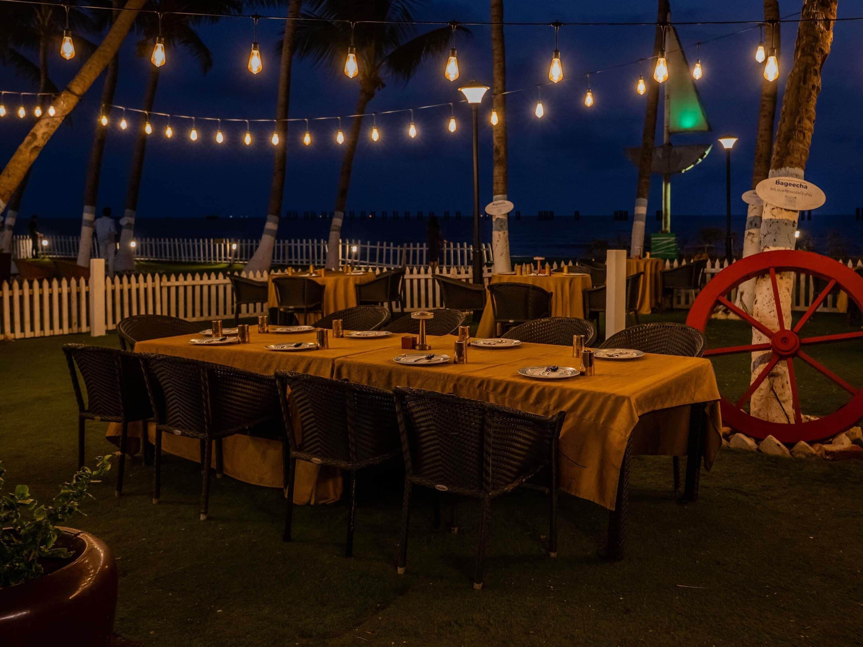 An outdoor dining area with tables set for two, surrounded by chairs, under string lights.