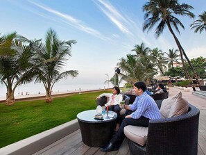 Eine Terrasse mit Korbmöbeln, einem Glastisch und Blick auf den Strand und Palmen.