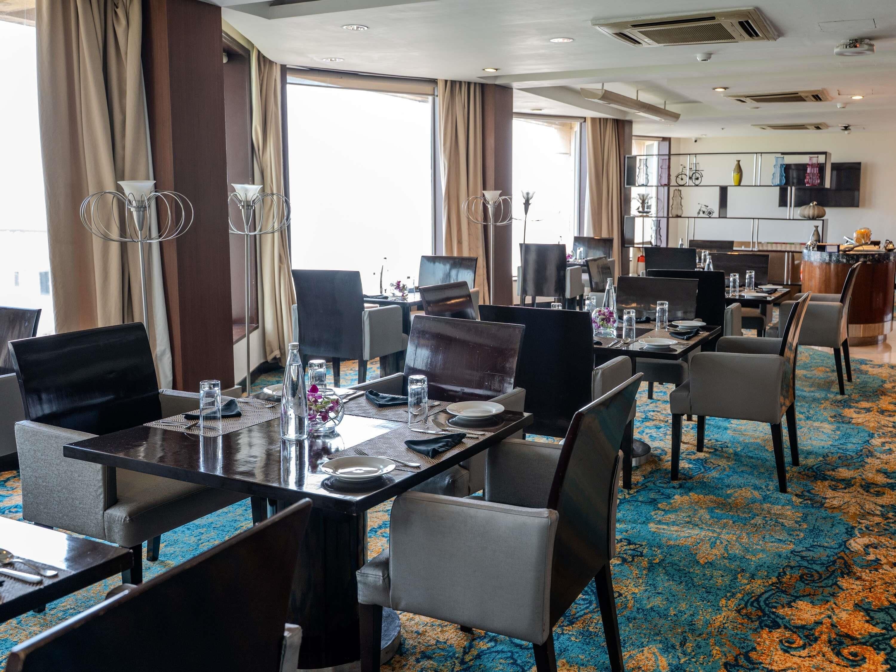 Une salle à manger d’hôtel avec des tables dressées pour les clients, une cuisine en arrière-plan et une vue sur la ville à travers de grandes fenêtres.