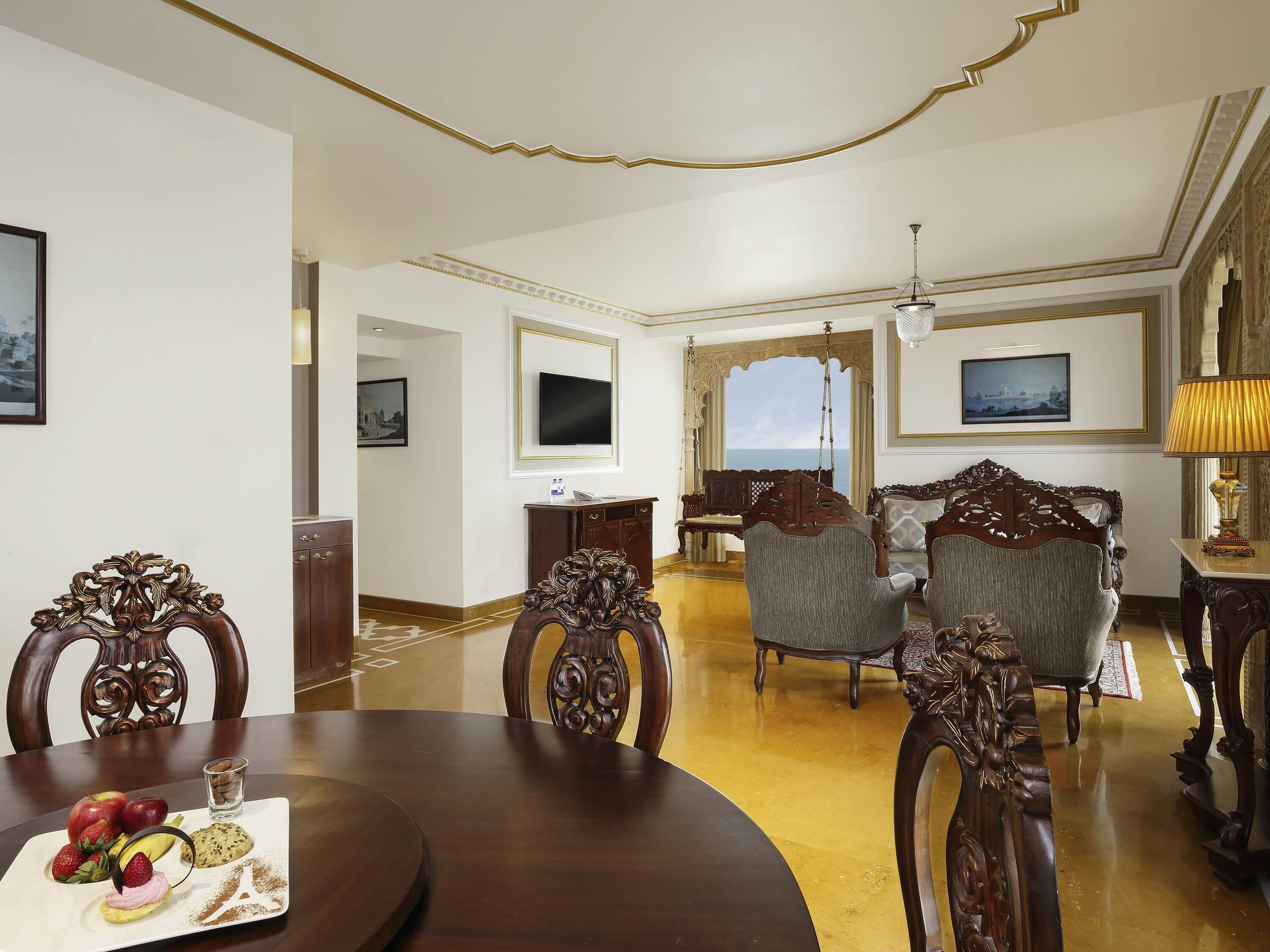 A spacious living room with ornate wooden furniture, a dining table with fruit and a glass of water, and a large window offering a view of the sea.