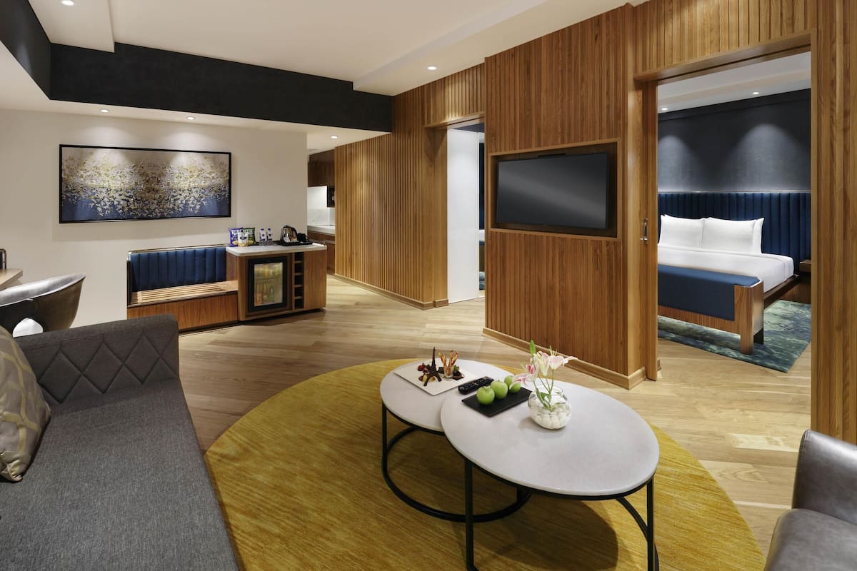 Habitació d'hotel moderna amb un sofà, una taula de centre rodona, una televisió i un llit amb capçalera blava.