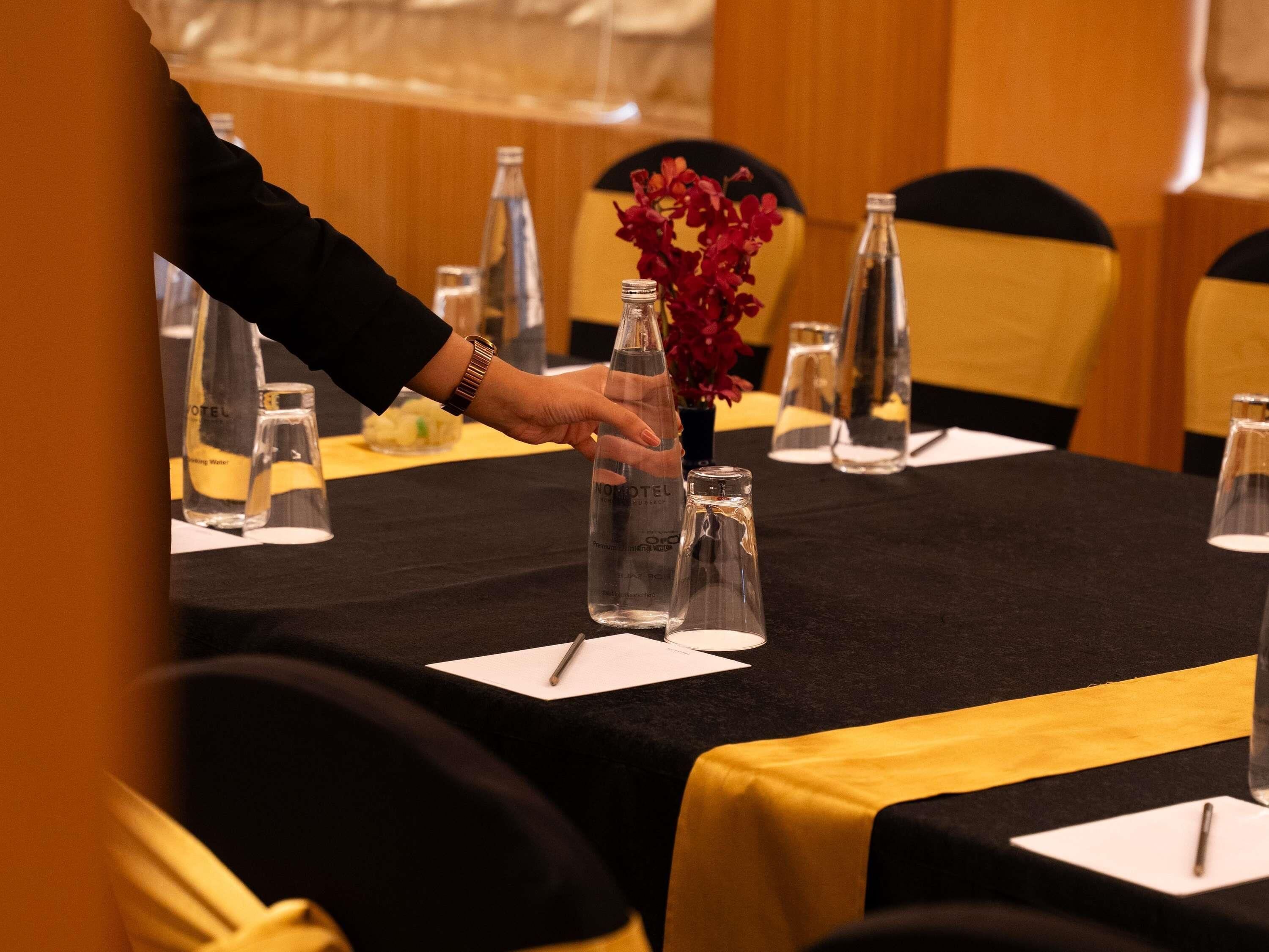 Una mesa de hotel decorada con botellas de vidrio transparente, un florero con flores rojas y una botella de agua.