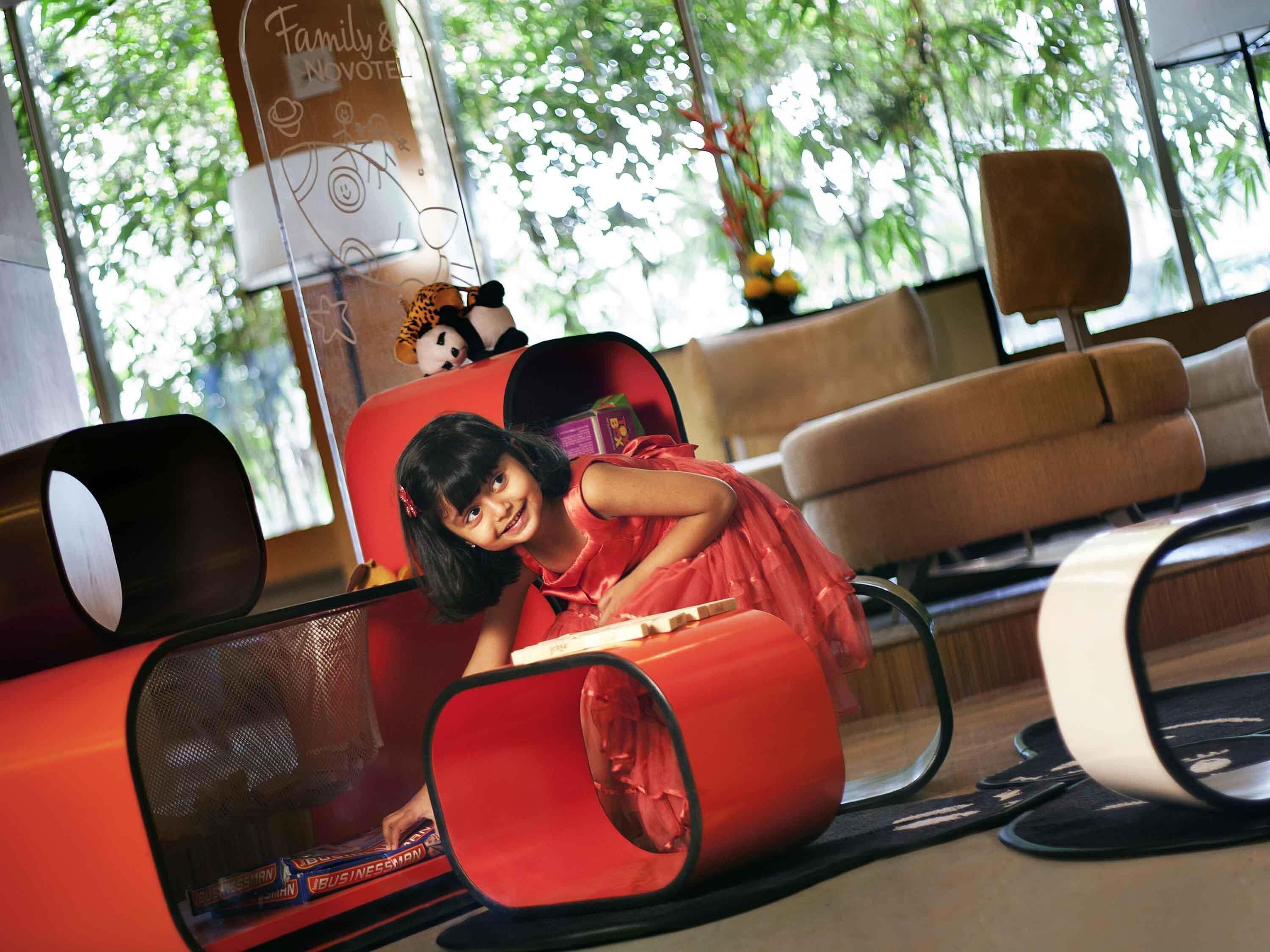A child in a red dress seated in a red chair with a toy panda on the backrest, in a room with large windows and a view of trees.