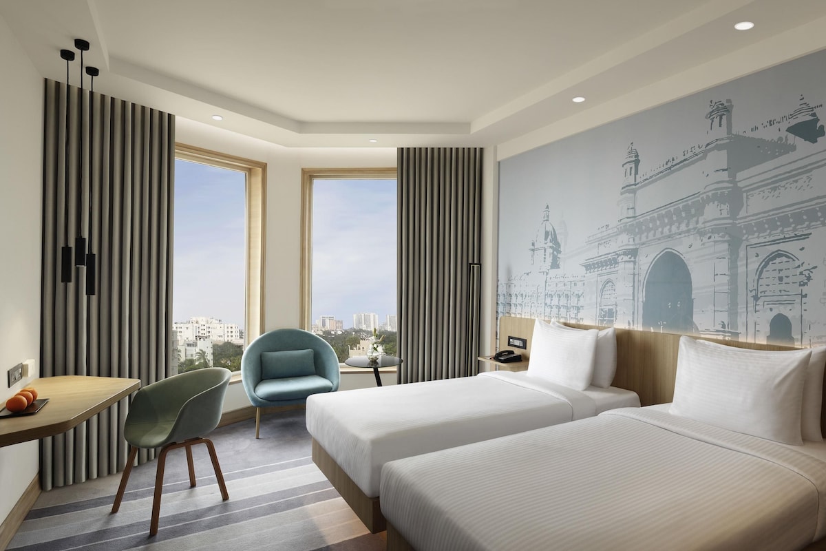 Ein Hotelzimmer mit zwei Betten, einem Schreibtisch, einem Stuhl, einem Nachttisch und einem großen Fenster mit Blick auf die Stadt.