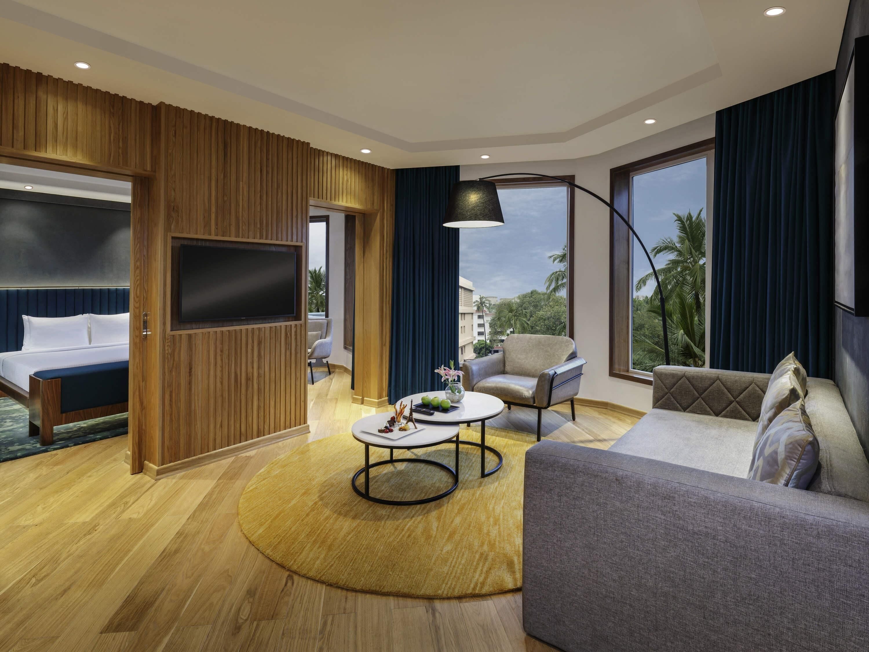 A modern hotel room with a bed, a sofa, a round coffee table, a TV, and large windows with a view of palm trees.