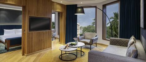 A modern hotel room with a bed, a sofa, a round coffee table, a TV, and large windows with a view of palm trees.