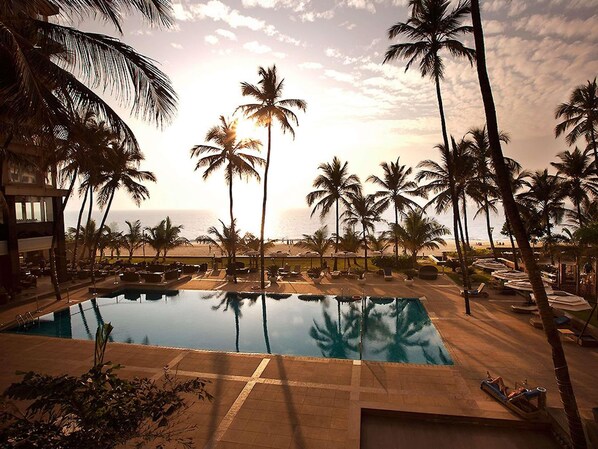 A resort pool area with palm trees, a clear sky, and a view of the ocean.