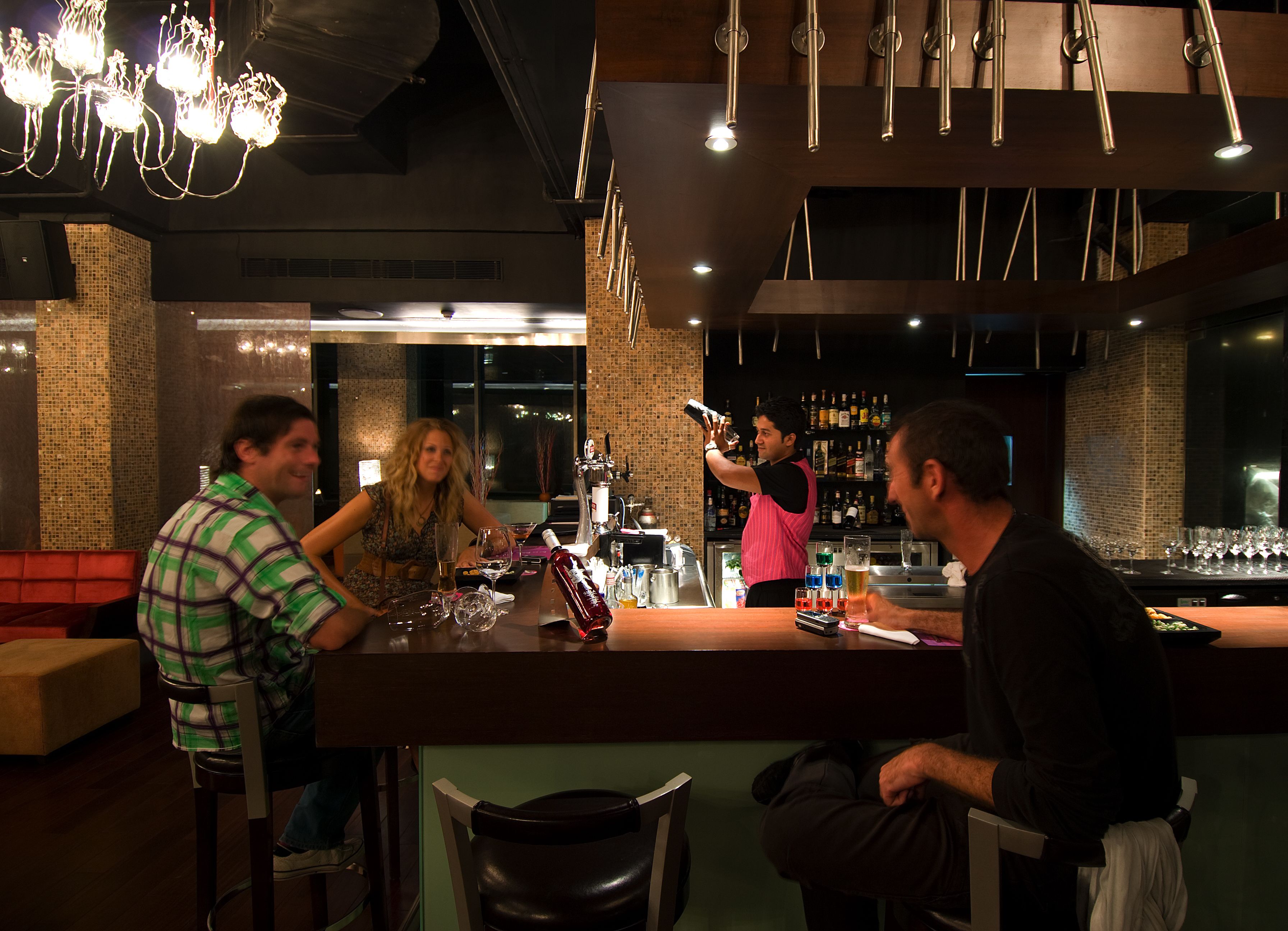 A bar area with a bartender preparing drinks, a seated customer, and another customer at a table.