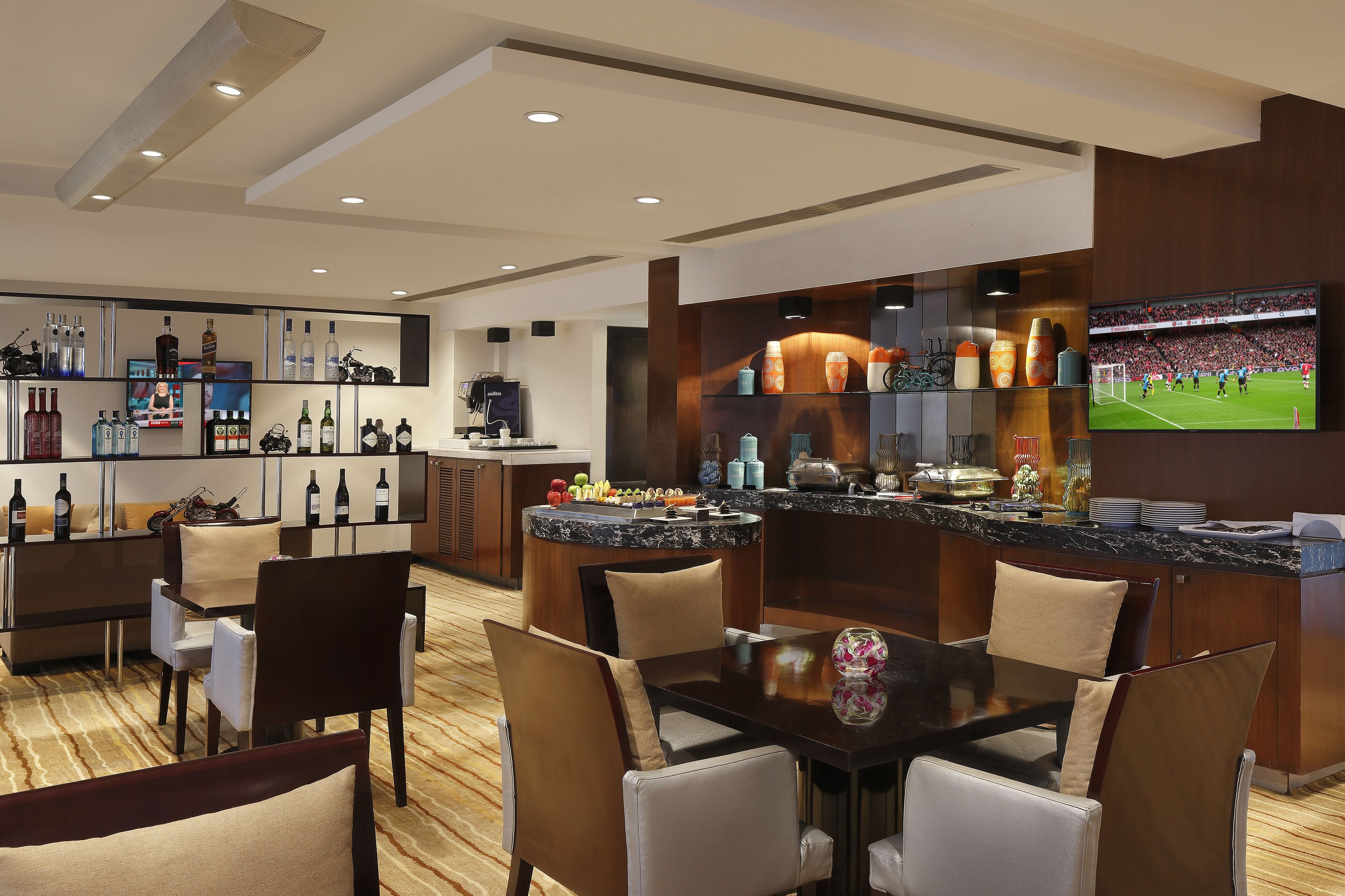 A modern hotel bar with a large TV screen, a variety of bottles, and a seating area with chairs and a table.