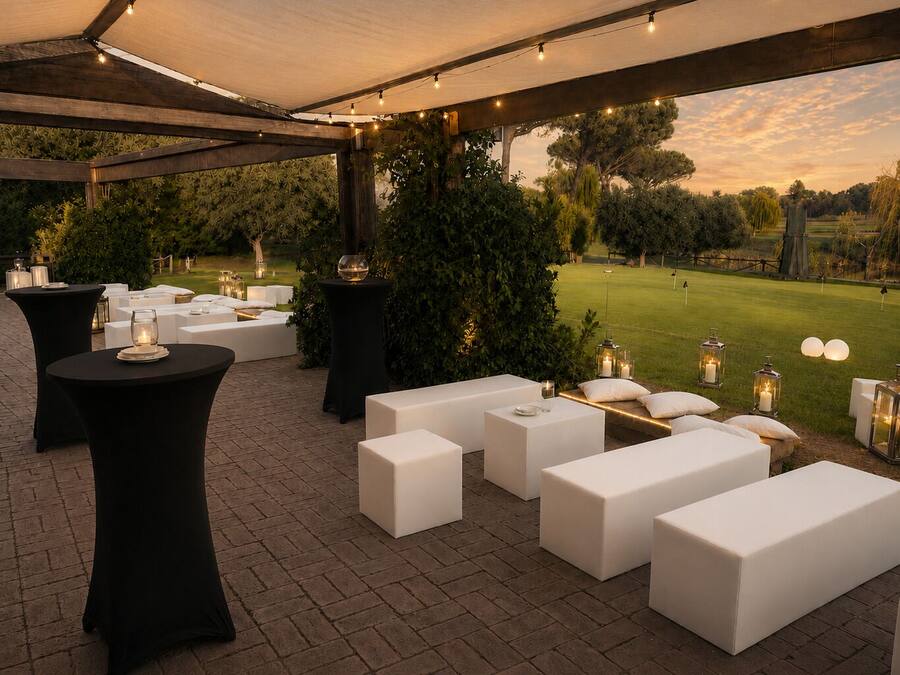 An outdoor seating area with round and rectangular white tables, black round tables, and white cushioned benches under a covered pergola with string lights.