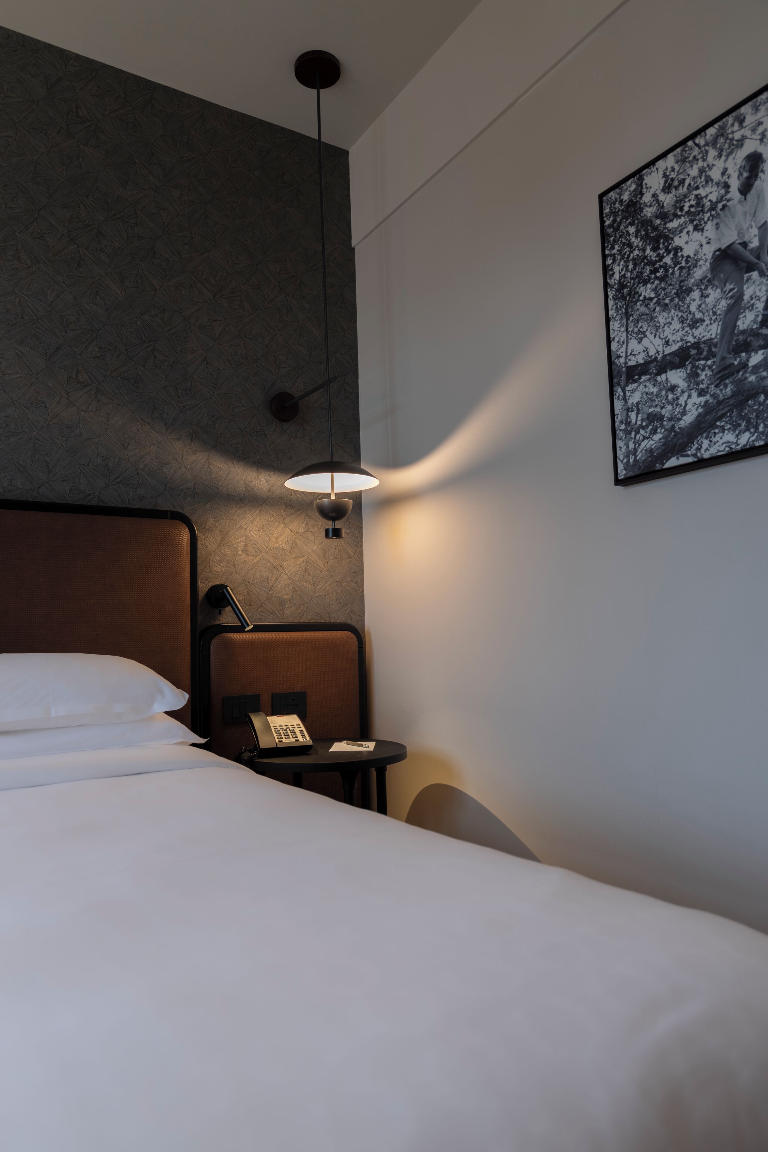 Ett hotellrum med en säng, ett sängbord med en telefon, en lampa och en tavla som hänger på väggen.