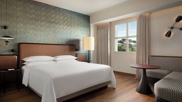 A hotel room with a bed, bedside tables, a lamp, a window with curtains, a sofa, and a small table.